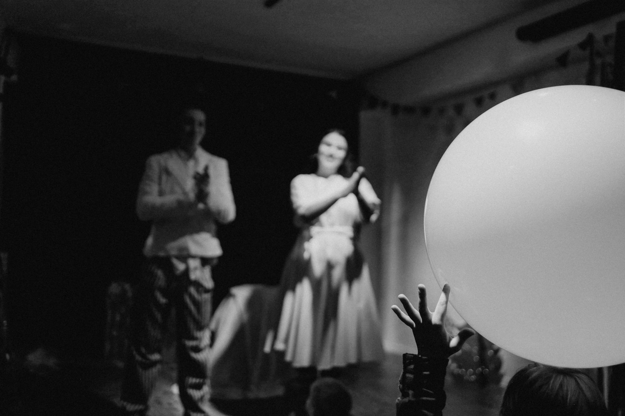 Monochrome photo of performers on stage with a child's hand reaching for a balloon.